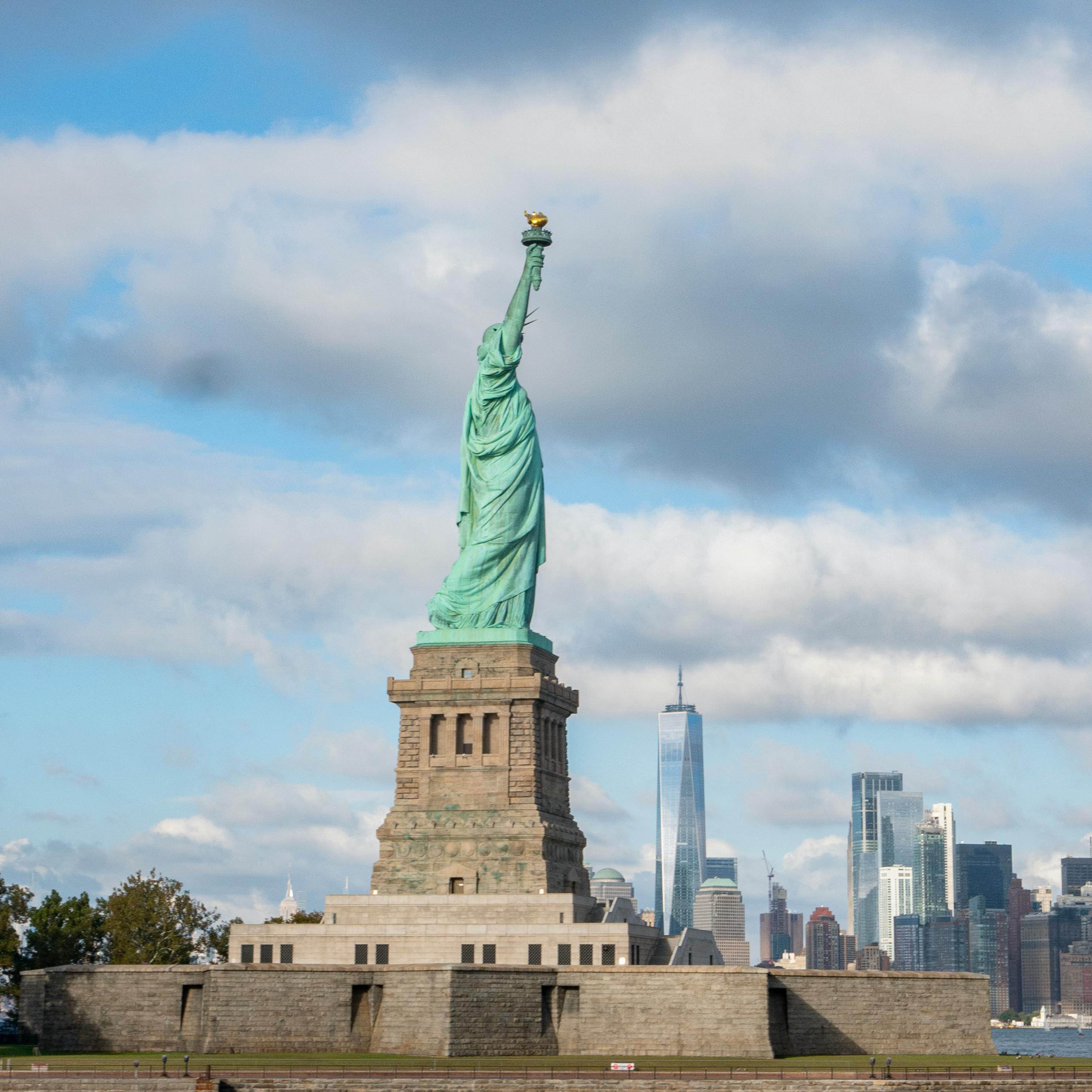 Iconic Statue of Liberty with New York City skyline in the background under a partly cloudy sky.